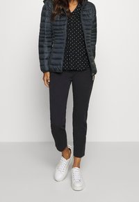 Giacca nera imbottita con cuciture orizzontali, camicetta nera a pois, pantaloni scuri e sneakers bianche con lacci e suole testurizzate.