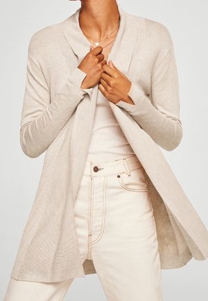 Beige cardigan met ribbelstructuur, open voorkant, lange mouwen en een sjawlboord, gedragen over een lichte top en gecombineerd metlichte spijkerbroek.