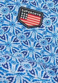 Tela azul con patrón que presenta un diseño floral, acompañada de un parche negro con "SUPERDRY" y un motivo de la bandera estadounidense.