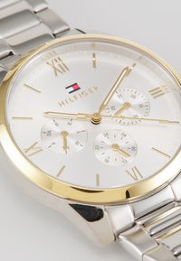 Reloj de plata y oro con una esfera redonda, tres subesferas, números romanos y una correa de metal. Presenta un diseño simple y elegante.