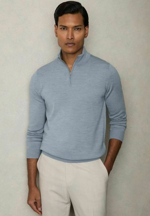 Pulover cu fermoar de culoare albastru deschis, dintr-un material moale și texturat, cu mâneci lungi, purtat peste pantaloni bej deschis. Design minimalist, fără modele.