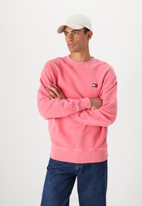 Felpa rosa con polsini e orlo a coste, caratterizzata da un piccolo logo ricamato. Abbinata a jeans blu scuro e un cappellino chiaro.