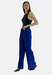Svart ärmlös bodysuit i kombination med flödande blå plisserade byxor med resårmidja och en texturerad yta. Svarta spetsiga skor.