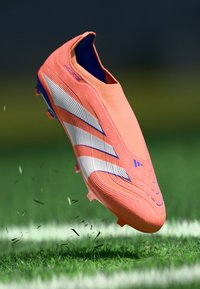 Bota de fútbol de un vibrante color naranja con un diseño aerodinámico, parte superior texturizada, rayas blancas y acentos azules. Elevada sobre la hierba, mostrando movimiento.