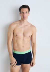 Boxer en coton bleu marine avec une taille verte arborant le logo "LACOSTE". Texture lisse, design ajusté, longueur mi-cuisse.