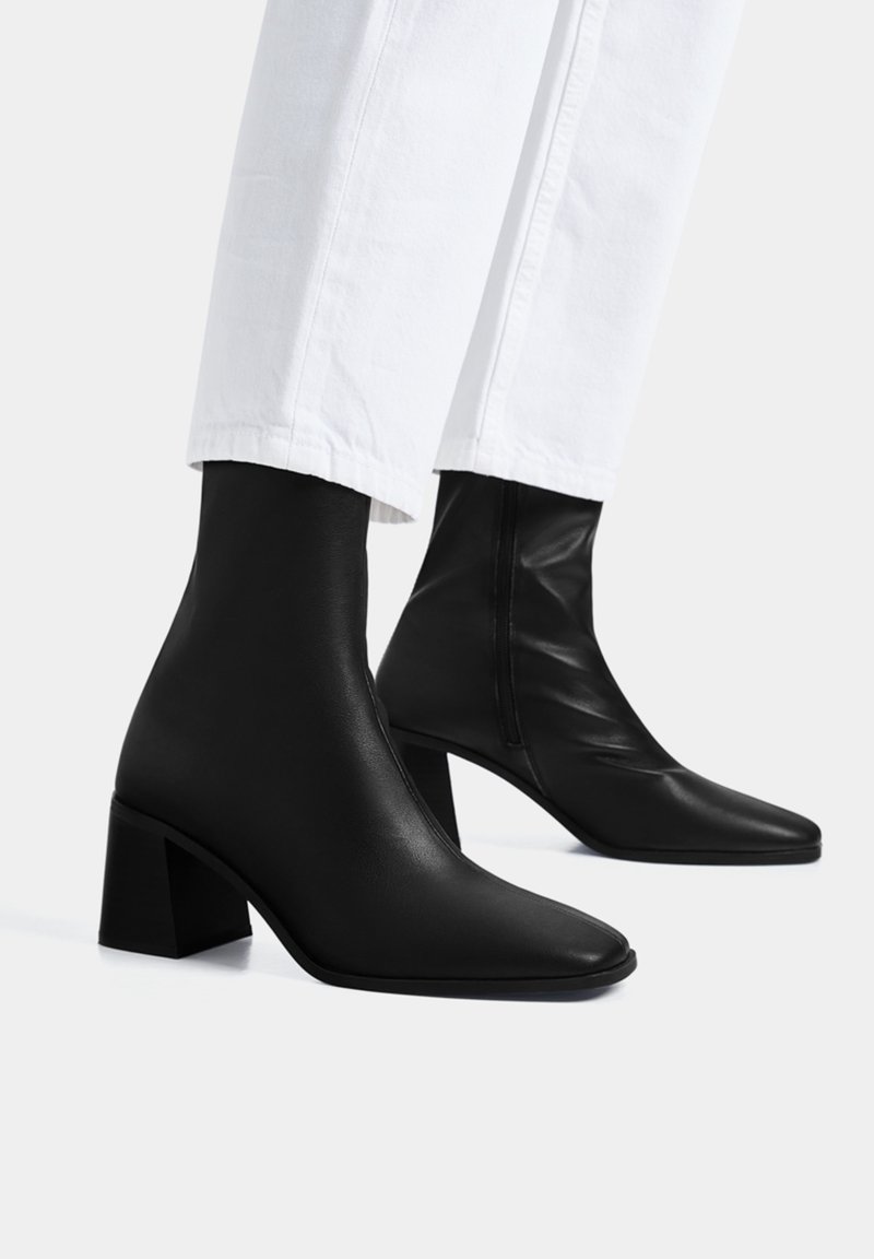 Bottes chevilles en cuir noir avec talons blocs portées avec un pantalon blanc, présentées sur un fond clair uni.