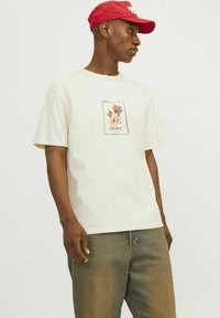 Beige kortærmet t-shirt med et blomster grafisk design og "atelier" tekst på forsiden, parret med lyse denim jeans og en rød kasket.