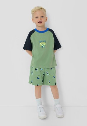 Blond chłopiec stoi, ubrany w zielono-czarną koszulkę z motywem piłkarskim oraz pasujące do niej spodenki, białe skarpetki i białe tenisówki.