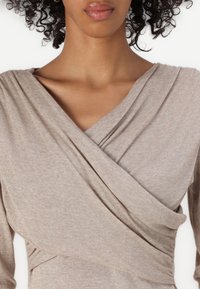Beige inneslutande topp gjord av mjukt tyg med draperad design och V-halsringning. Har långa ärmar och rynkad detalj vid midjan.