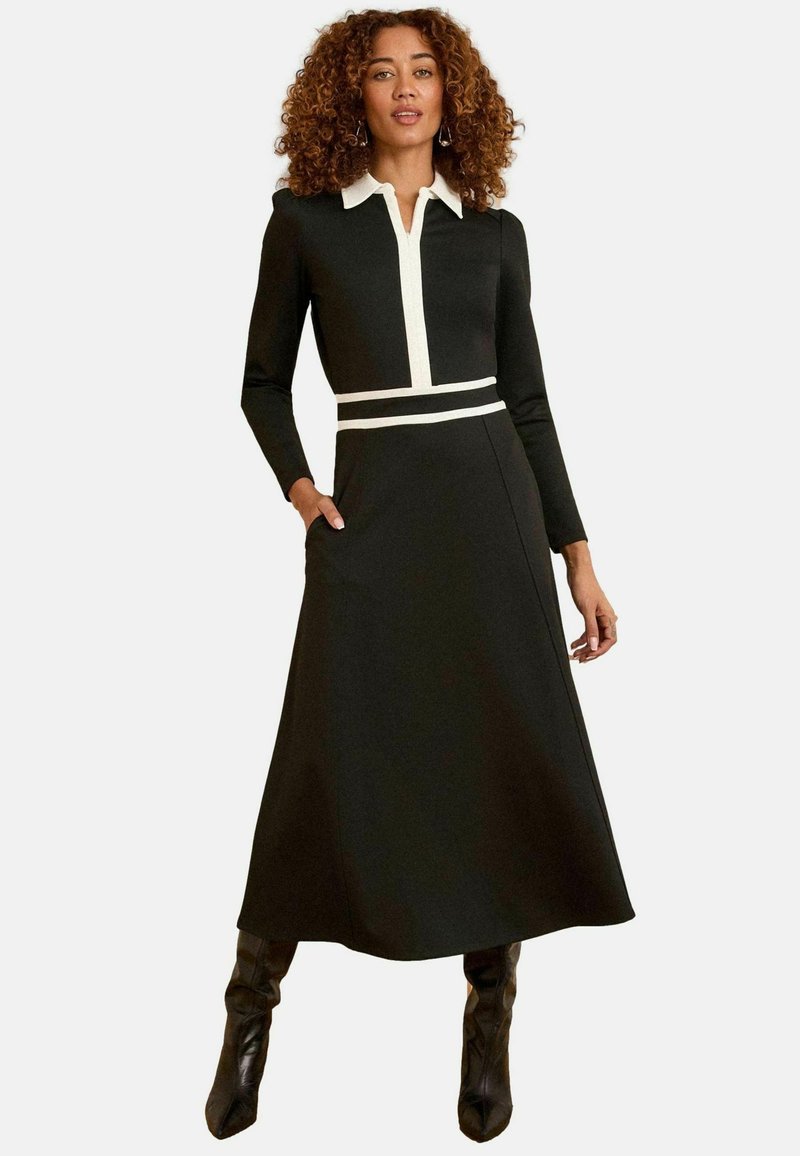 Vestido negro de manga larga con cuello blanco y cinturilla a rayas, con un corsé entallado, falda acampanada y bolsillos laterales. Textura suave.