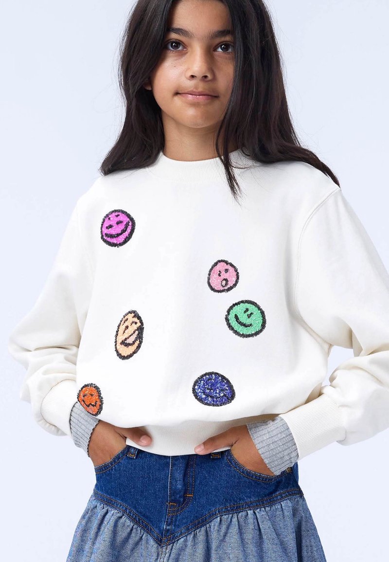 Biela mikina s farebnými usmievavými emoji rôznych veľkostí; má rebrované manžety a okrúhly výstrih. Skombinovaná s džínsovou sukňou.