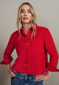 Camicia in corduroy rosso con bottone frontale, taschino singolo sul petto e maniche arrotolate, abbinata a jeans in denim blu. Tessuto liscio e design a costine.