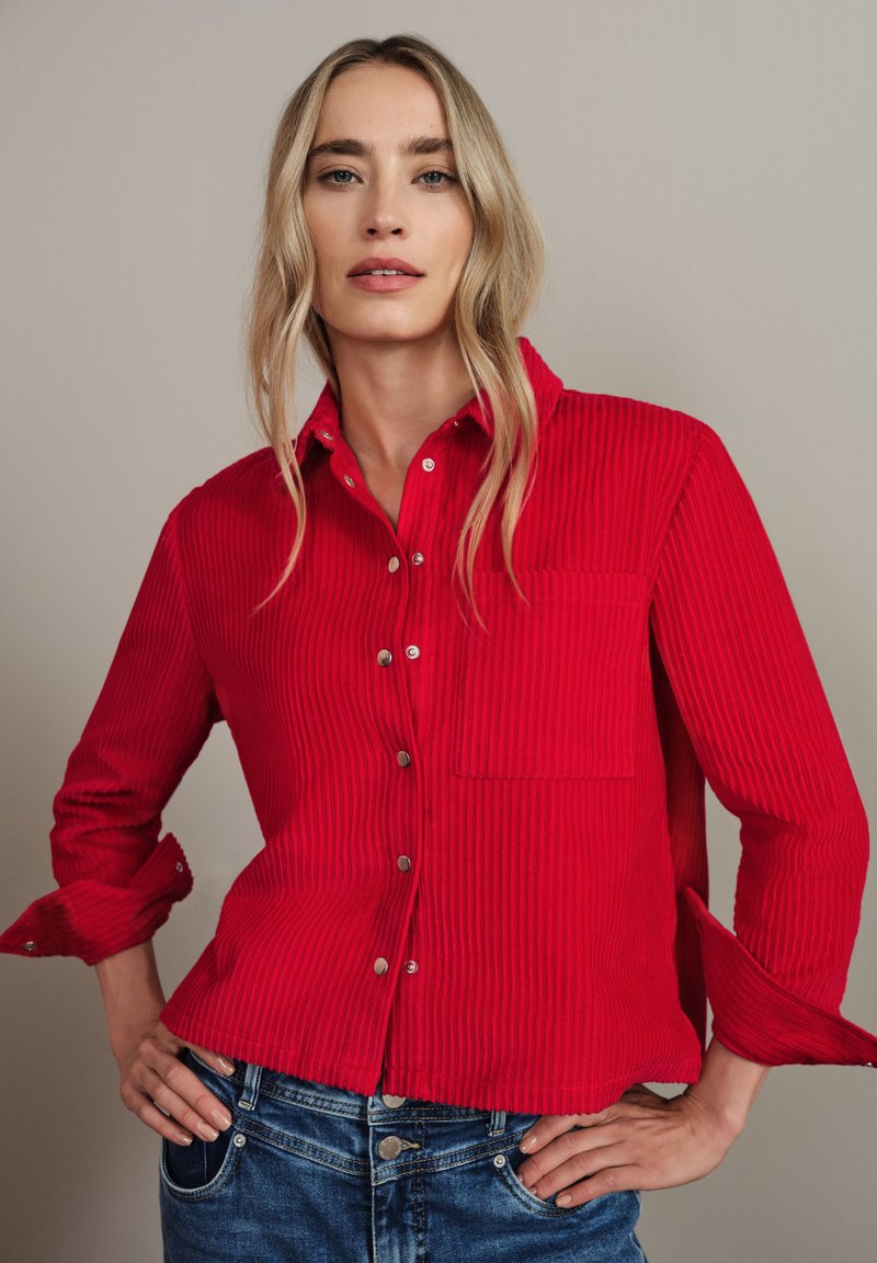 Camicia in corduroy rosso con bottone frontale, taschino singolo sul petto e maniche arrotolate, abbinata a jeans in denim blu. Tessuto liscio e design a costine.