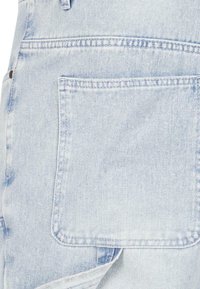Gai zilgani denim kombinezons ar lielu aizmugurējo kabatu, kas nostiprināta ar stiprinājumiem, sānu siksnu un klasisku brīvu piegriezumu.
