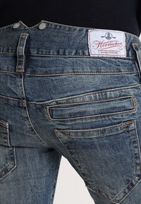 Mörkblå denimjeans med en texturerad yta, flera bakfickor med sömnadsdetaljer och en synlig logotyp på midjebandet.