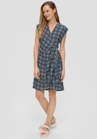 Navyblauwe wikkeljurk met een bloemenpatroon, korte capmouwen en een geraffineerde taille. Gecombineerd met lichtgekleurde, gevlochten plateausandalen.