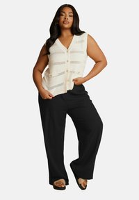 Stickad krämfärgad väst med v-ringning, öppen vävstruktur och knappstängning, parat med vida svarta byxor och sandaler.