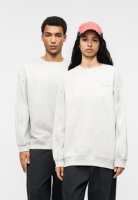 Ανοιχτό γκρι φούτερ με μακριά μανίκια διαθέτουν πλαϊνές φερμουάρ. Το φόντο είναι απλό, αναδεικνύοντας τα ρούχα που φορούν δύο άτομα.