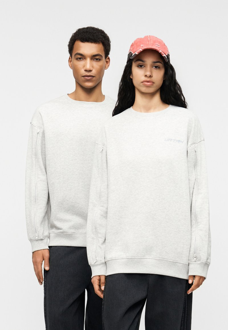 Ανοιχτό γκρι φούτερ με μακριά μανίκια διαθέτουν πλαϊνές φερμουάρ. Το φόντο είναι απλό, αναδεικνύοντας τα ρούχα που φορούν δύο άτομα.