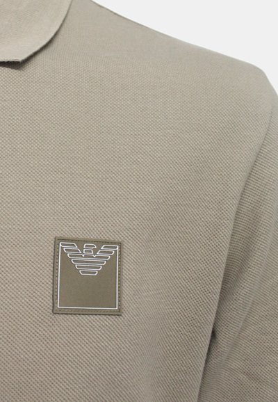 Bež polo majica z teksturirano tkanino. Ima kvadratni emblem v zgornjem levem kotu z stiliziranim logotipom v belih in sivih odtenkih.