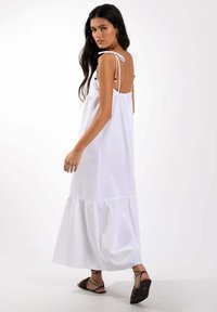 Vestido maxi blanco con una tela texturada, tirantes delgados y un dobladillo en capas. Presenta un diseño de espalda descubierta y se combina con sandalias negras de tiras.