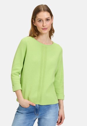 Maglione verde chiaro, texture a coste, con scollatura rotonda e maniche tre-quarti; presenta linee verticali al centro e polsini arrotolati.