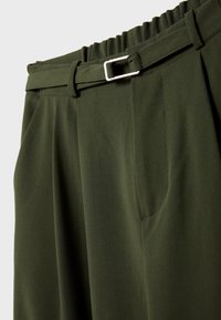 Pantalon vert foncé avec devant plat, plis, finition texturée, taille élastique et ceinture avec boucle argentée.