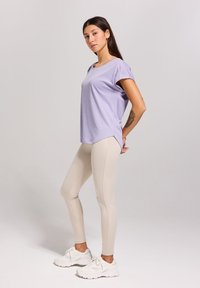 Licht paarse sport-T-shirt met kapmouwen, gecombineerd met beige leggings. De outfit wordt aangevuld met witte sportschoenen.