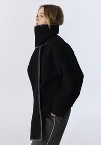 Abrigo de lana negro con un cuello alto y costuras claras en contraste, con un ajuste relajado y bolsillos laterales, combinado con pantalones grises.