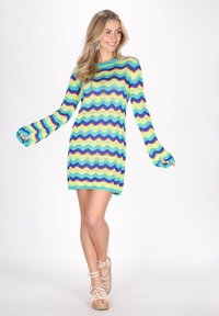 Gebreide jurk in teal, limoen en roze golvende strepen. Losse mouwen, ronde hals, open stof en korte lengte. Draagt met beige gespenhakken.