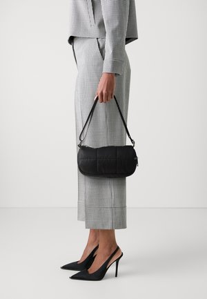 Sac à main matelassé noir avec une forme rectangulaire slim, tenu par une main. Les tenues présentent un motif à carreaux noir et blanc, avec des talons aiguilles.