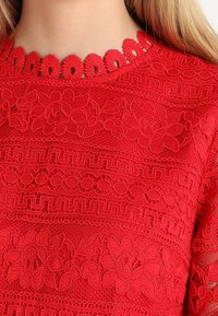 Blouse rouge brodée présentant des motifs floraux et géométriques complexes, avec une finition texturée et un col à volants.