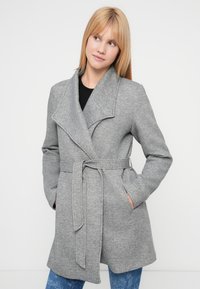 Γκρι παλτό τύπου wrap με μεγάλο γιακά, ζώνη στη μέση, μακριά μανίκια, τσέπες, κατασκευασμένο από μαλακό υφασμάτινο υλικό, μπροστά από μια απλή λευκή επιφάνεια.