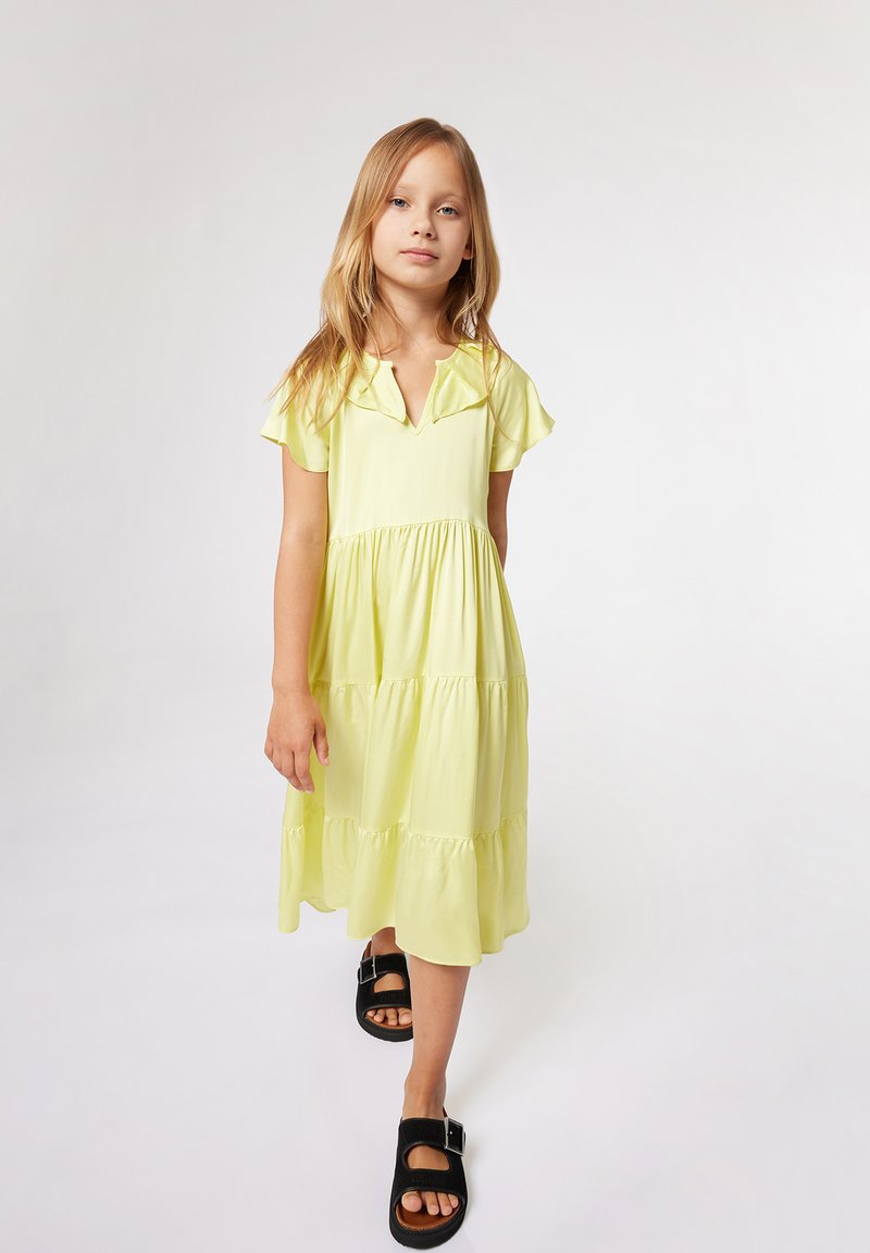 Robe jaune à volants avec des manches flottantes, décolleté en V et texture lisse. Associée à des sandales noires avec des sangles à boucle.
