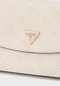Cremefarbene Guess-Handtasche aus strukturiertem Leder, mit geprägten Mustern und einem goldfarbenen, dreieckigen Logoakzent auf der Vorderseite.