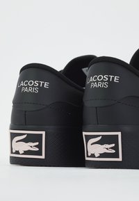Juodi sportbačiai su tekstūruotu guminiu padu, kuriuose yra baltas užrašas "LACOSTE PARIS" ir rožinis krokodilo logotipas ant kulno. Lygus paviršius.