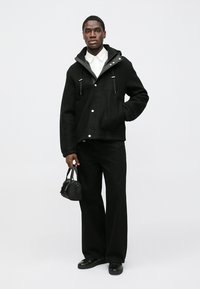 Sort jakke med hætte, trykknapper og lange ærmer. Kombineret med sorte bukser med vide ben og bærer en lille sort taske. Ensfarvet baggrund.