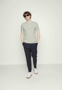 Jonge man in een gestreepte polo, donkere broek, witte sneakers en zonnebril, staande met zijn handen in zijn zakken tegen een effen achtergrond.