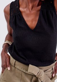 Haut noir sans manches avec col en V, tissu texturé et accents en dentelle sur les épaules, associé à un pantalon kaki taille haute avec ceinture nouée.