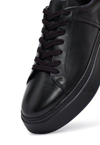 Zapatilla de cuero negra con suela texturizada gruesa y cordones negros, con costuras detalladas a lo largo del costado, angular sobre fondo blanco.