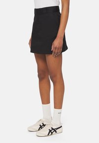 Černá sukně s upnutým pasem, předními kapsami a rovnou linií, kombinována s bílými ponožkami a béžovými teniskami s černými akcenty.