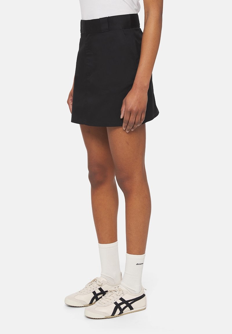 Černá sukně s upnutým pasem, předními kapsami a rovnou linií, kombinována s bílými ponožkami a béžovými teniskami s černými akcenty.