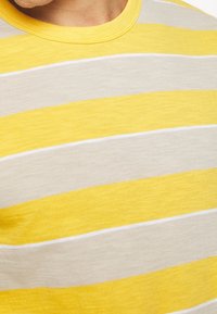 Tela a rayas amarilla y gris con una textura suave. Cuello redondo y mangas cortas. Presenta rayas horizontales llamativas y líneas blancas en contraste.