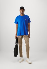 Uomo che indossa una maglietta blu, pantaloni beige, sneakers bianche e un cappellino nero, mentre tiene una borsa tote trapuntata nera contro uno sfondo grigio uniforme.
