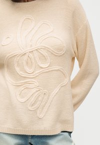 Μπεζ πλεκτό πουλόβερ με ανάγλυφο υφασμάτινο αφηρημένο floral σχέδιο φορεμένο με ανοιχτό μπλε τζιν.