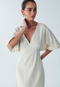 Helles beiges Leinenkleid mit tiefem V-Ausschnitt, kurzen Puffärmeln und tailliertem Oberteil. Glatte Textur, minimalistisches Design und keine sichtbaren Muster.