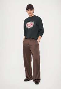 Ciemnozielona bluza z nadrukiem w kolorach czerwonym i białym, zestawiona z luźnymi, błyszczącymi brązowymi spodniami; model nosi brązową czapkę.