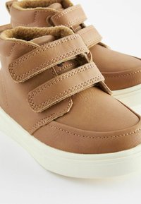 Bottes de cheville en cuir marron avec deux bandes Velcro, semelle en caoutchouc blanc et doublure en texture douce. Comprend des coutures le long des bords.