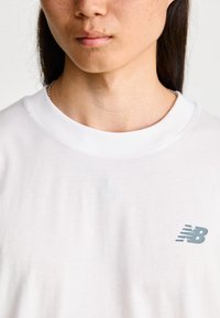 Bílý bavlněný tričko s krátkým rukávem a kulatým výstřihem. Obsahuje malé šedé logo "NB" na levé straně hrudi.
