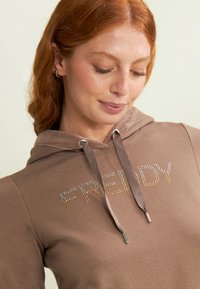 Felpa marrone con lettering di strass che scrive "FREDDY". Ha un cappuccio con coulisse e dettagli argentati nei passanti. Tessuto dalla consistenza liscia.